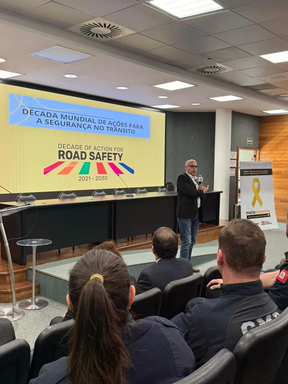 Ricardo Alves da Silva palestrando em palco com telão da Década Mundial de Ações para a Segurança no Trânsito